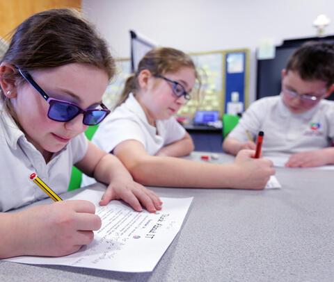 Two girls and a boy all wearing glasses sitting at a table writing with pens at school