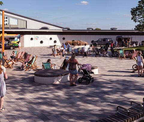 Ermine Street Church Academy School Plaza. It's a sunny day and members of the community are walking and relaxing in deckchairs in front of the school building