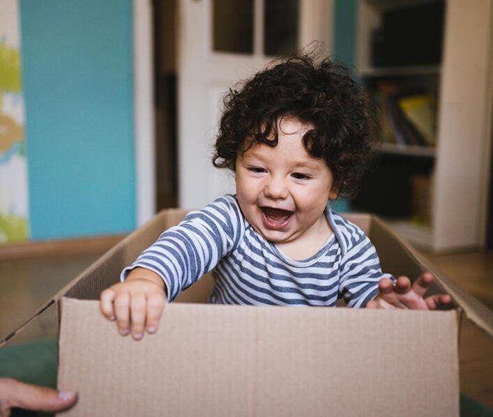 A toddler with dark curly hair plays in a packing box